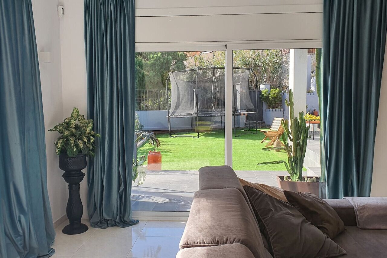 Un salon avec une porte-fenêtre coulissante donnant sur un jardin arrière comprenant une pelouse, un trampoline et des plantes en pot.