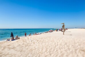 Een zandstrand met mensen die zonnebaden en zwemmen, een redershut en een helderblauwe lucht.
