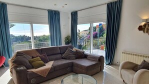 A modern living room with a sectional sofa, a glass coffee table, and large windows offering a view of the outdoors. - Opening Doors Premià Dalt Amazing views with pool (Premià de Dalt)