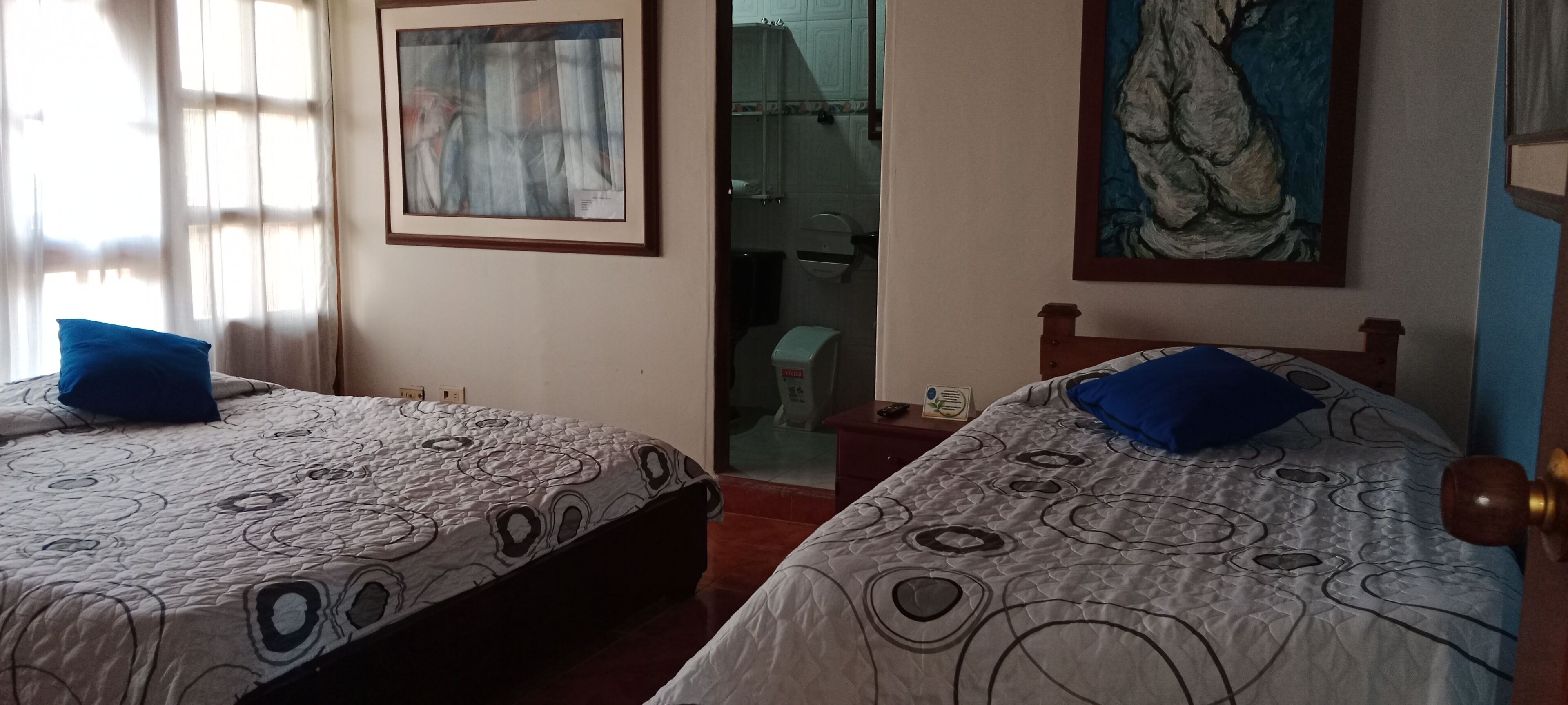 Two beds with patterned bedding and blue pillows, a framed abstract painting, and a bathroom visible through a glass door.