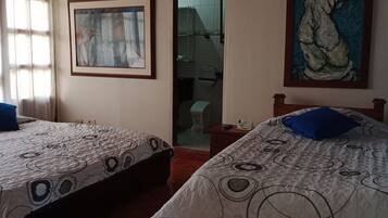 Two beds with patterned bedding and blue pillows, a framed abstract painting, and a bathroom visible through a glass door.