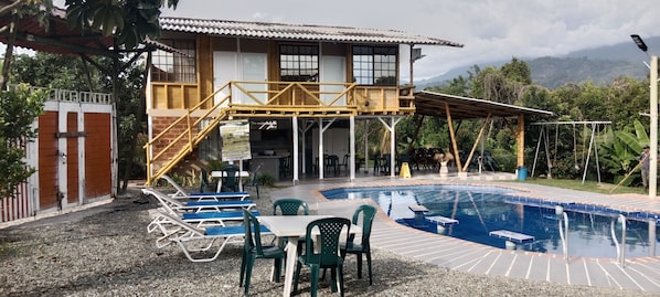 Una casa de dos pisos con piscina, mobiliario de exterior y una escalera que lleva a un balcón.