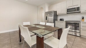 Una cocina moderna con una mesa de comedor de vidrio, sillas blancas, electrodomésticos de acero inoxidable y piso de baldosas.