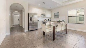 Una cocina moderna con electrodomésticos de acero inoxidable, un comedor con mesa de vidrio y sillas blancas, y un pasillo que conduce a una puerta.