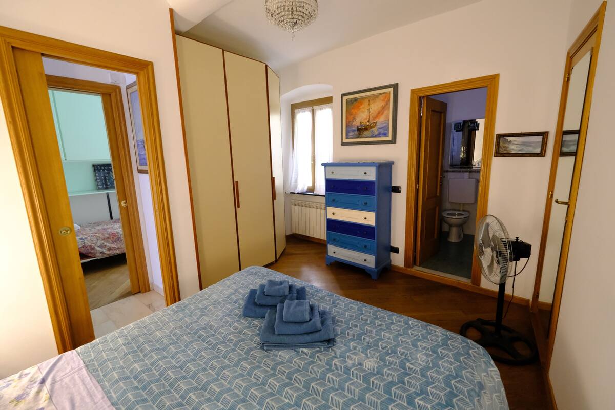 A bedroom with a bed, a wardrobe, a dresser, a fan, and a painting on the wall.