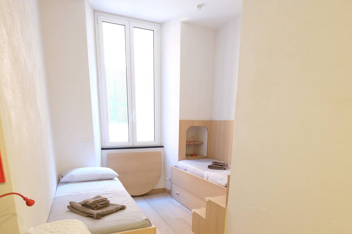 A compact bedroom with two beds, a small desk, and a window providing natural light.