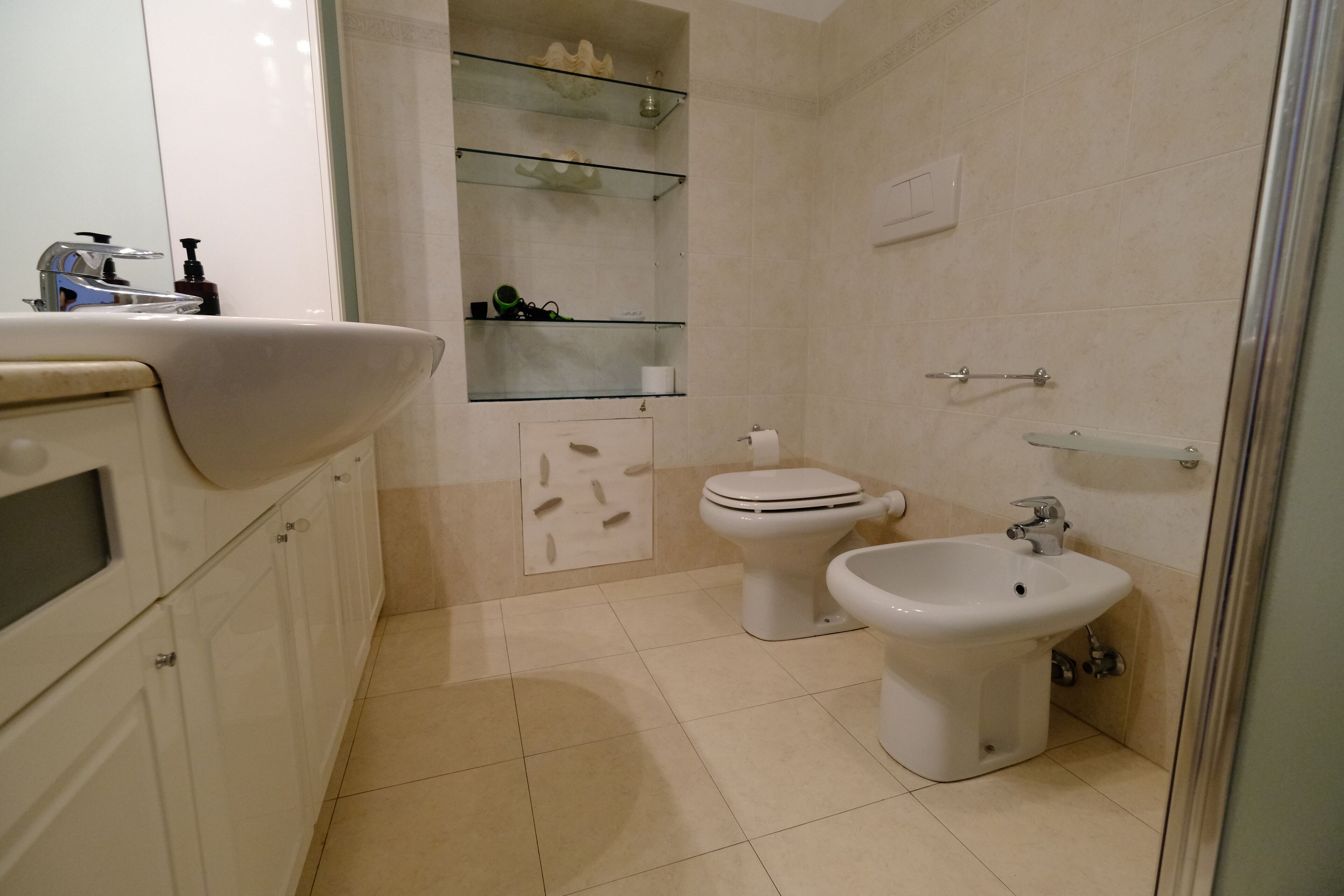 A modern bathroom with a white sink, toilet, and bidet, glass shelves, and a tiled floor.