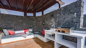 Un muro de piedra, un techo de madera y un banco blanco con cojines rojos.