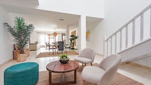Amplia sala de estar con una mesa de centro redonda de madera, un otomano azul turquesa y un sillón color beige. Al fondo, se observa un comedor con una mesa y sillas.