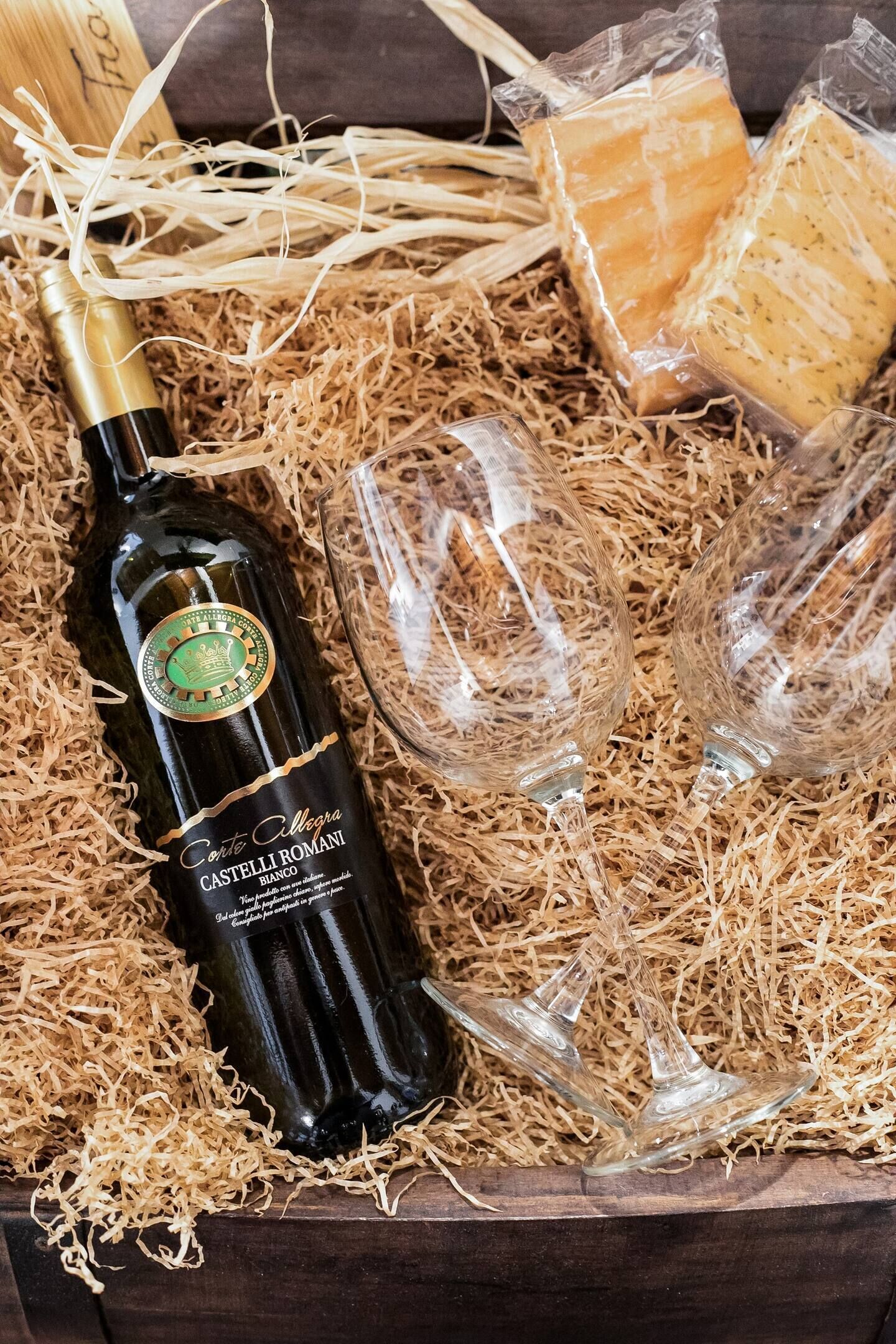 Una botella de vino de Castelli Romani, dos copas y queso envuelto en plástico, todo sobre un mantel de paja.