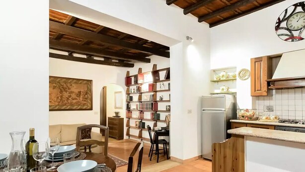 A dining area with a table set for two, a bookshelf, and a kitchen with a refrigerator and stove. - La Trasteverina apartment (Roma)