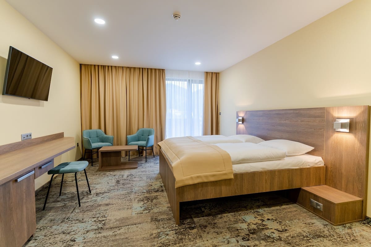Ein Hotelzimmer mit Bett, Schreibtisch, zwei Stühlen, Fernseher und einem Fenster mit Vorhängen.