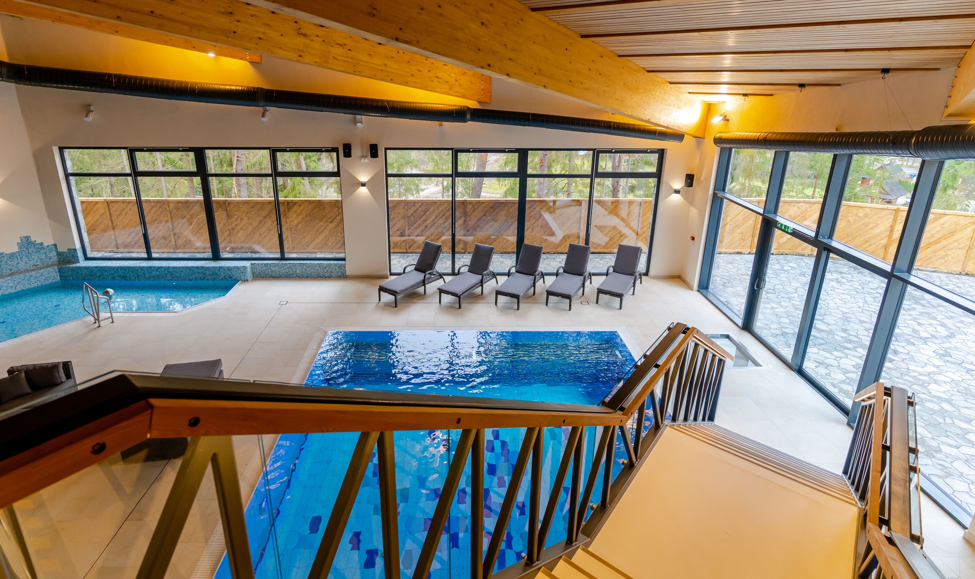 Una piscina interior moderna con vistas claras del entorno, techo de madera y zona de estar con sillones reclinables.