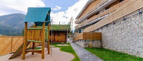 A playground with a climbing structure and a stone pathway leading to a building with a stone facade and wooden balconies.