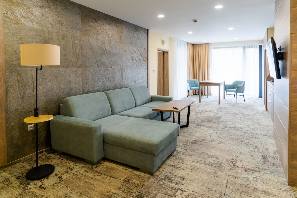 Ein modernes Hotelzimmer mit einer Eckcouch, einem kleinen Tisch, einer Stehlampe und einem an der Wand befestigten Fernseher.