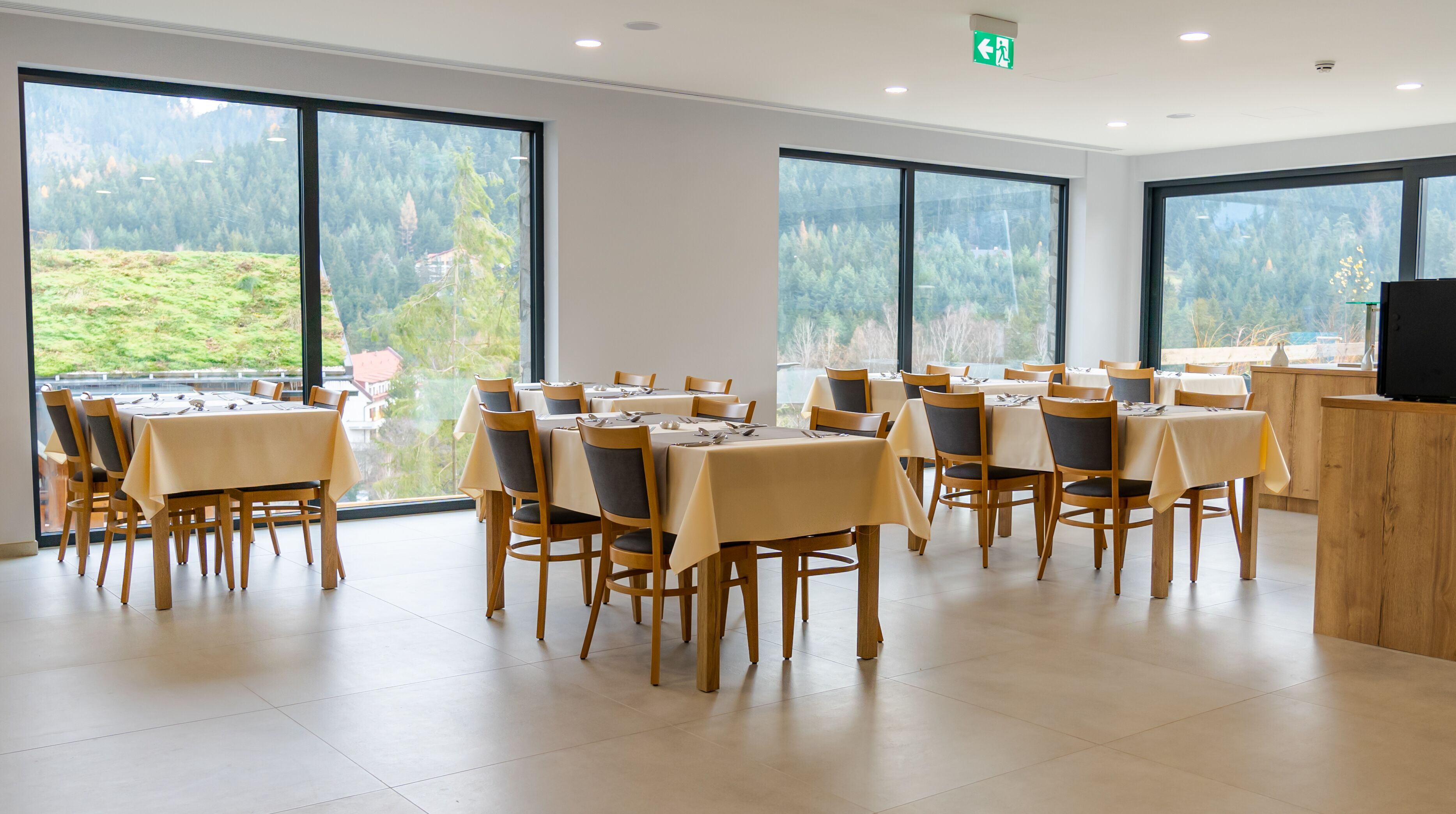A dining area with multiple tables and chairs, large windows with a view of a forested area, and a flat-screen TV on a wooden counter.