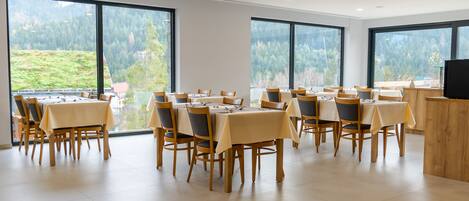 A dining area with multiple tables and chairs, large windows with a view of a forested area, and a flat-screen TV on a wooden counter.