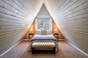 A bedroom with a sloped ceiling, a bed with a bench, two bedside tables, and a window with curtains.