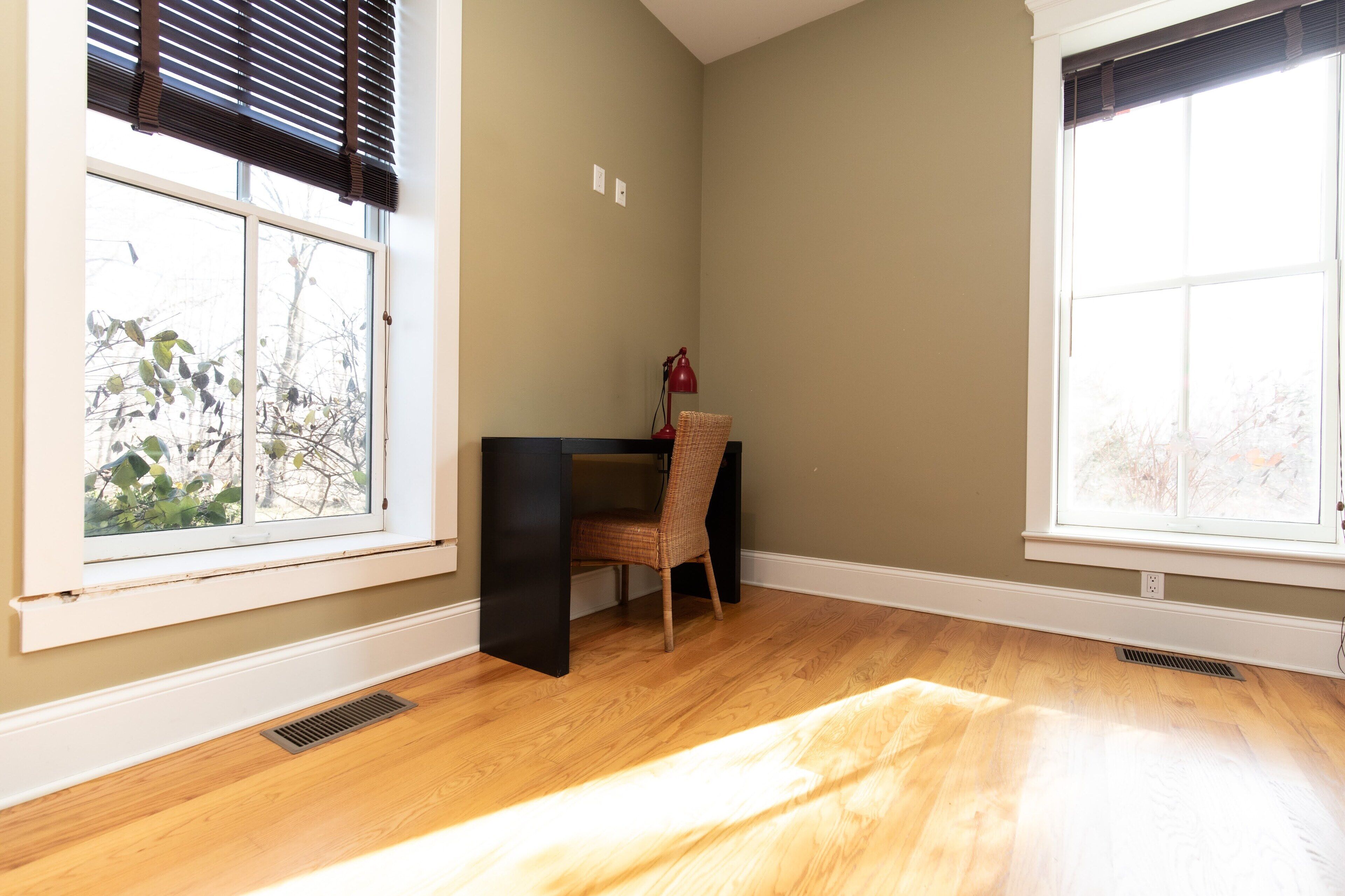A room with wooden flooring, a desk with a chair, and two windows with blinds.