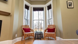 A corner room with two wicker chairs, a small table, and a bay window with a view of trees. - Furness Mansion (Chesterton)