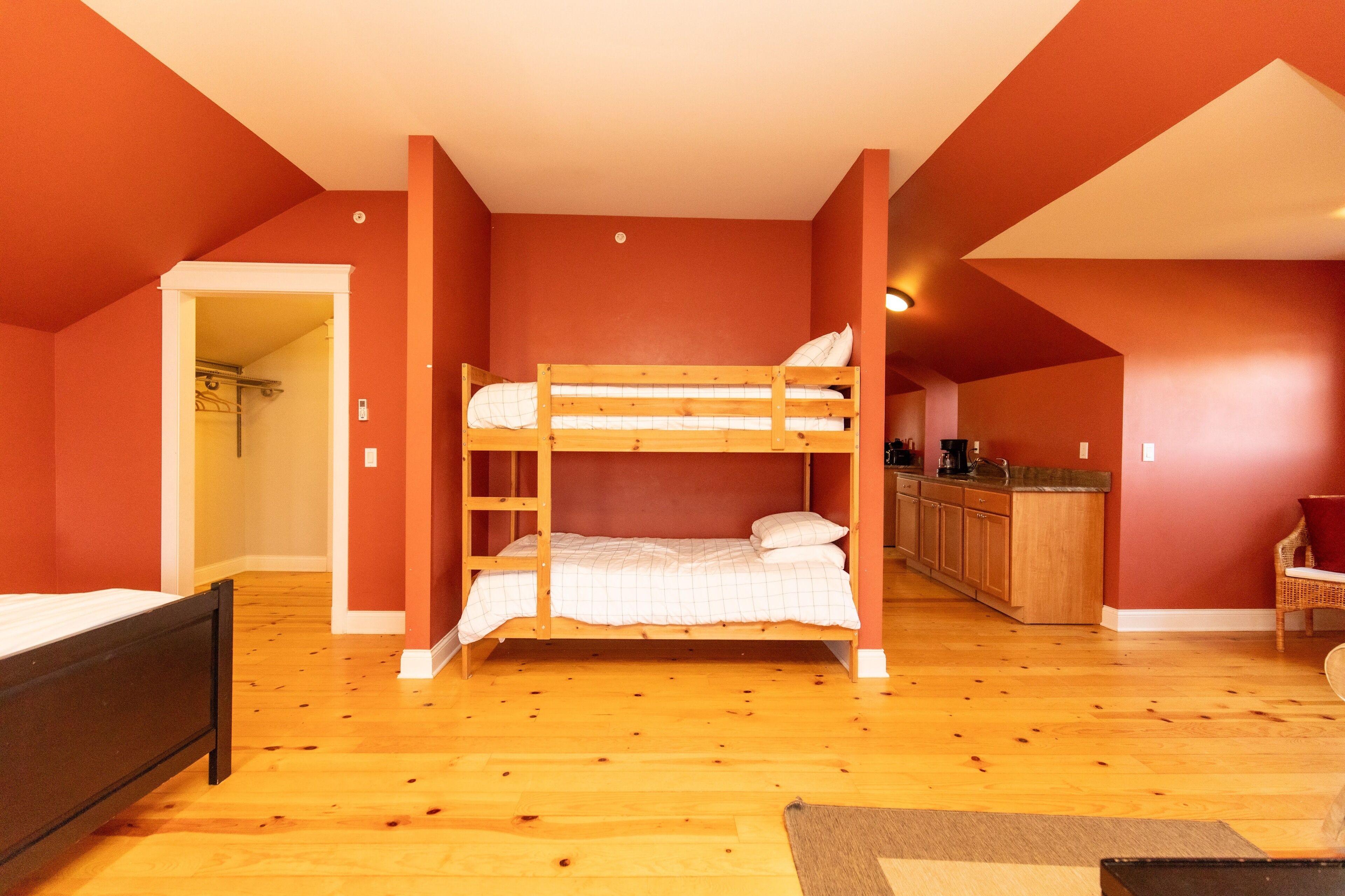 A room with a bunk bed, wooden flooring, a kitchen area, and a doorway leading to another room.