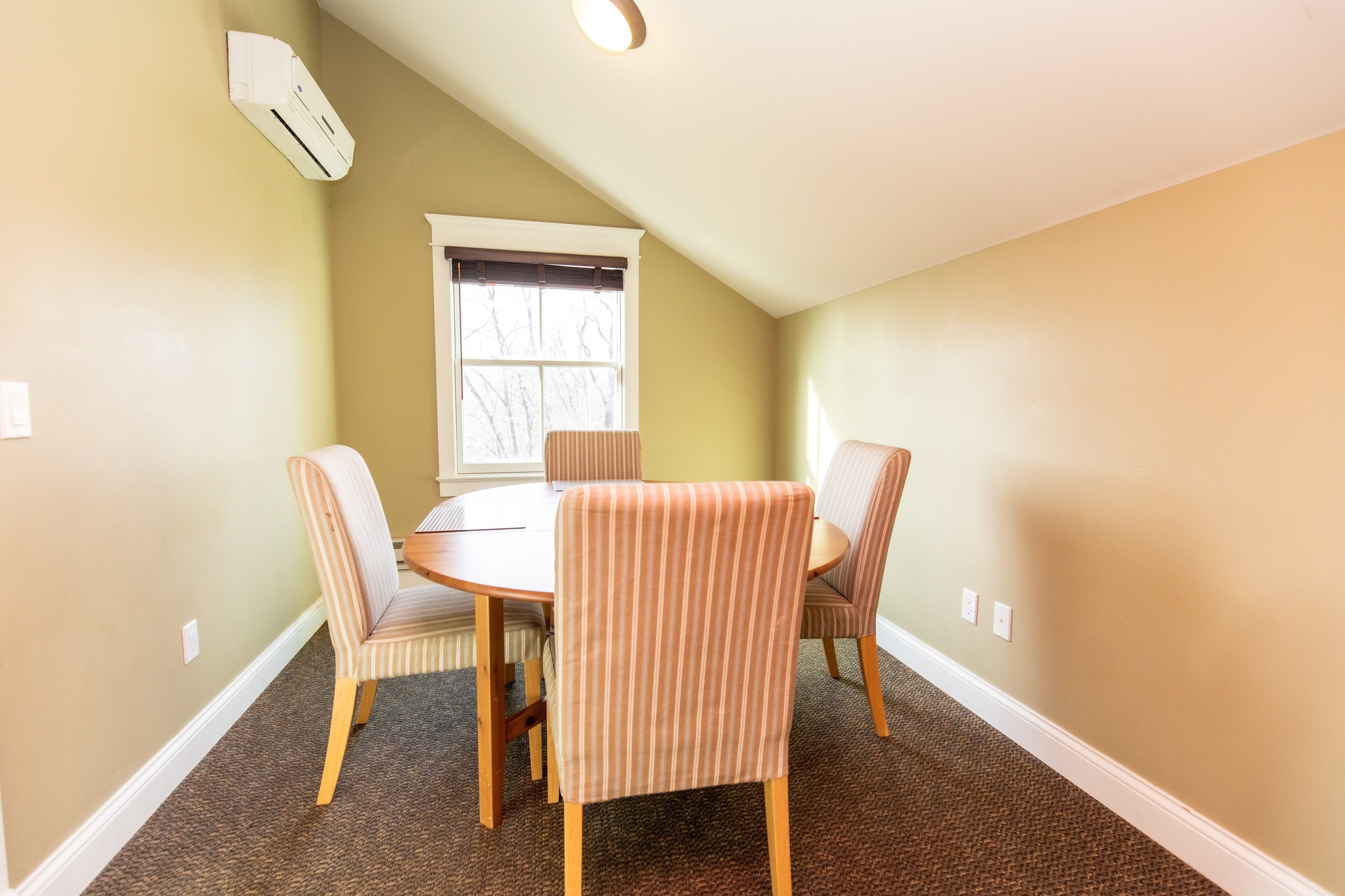 Un coin repas avec une table ronde et quatre chaises à rayures, une fenêtre avec des stores et un climatiseur fixé au mur.