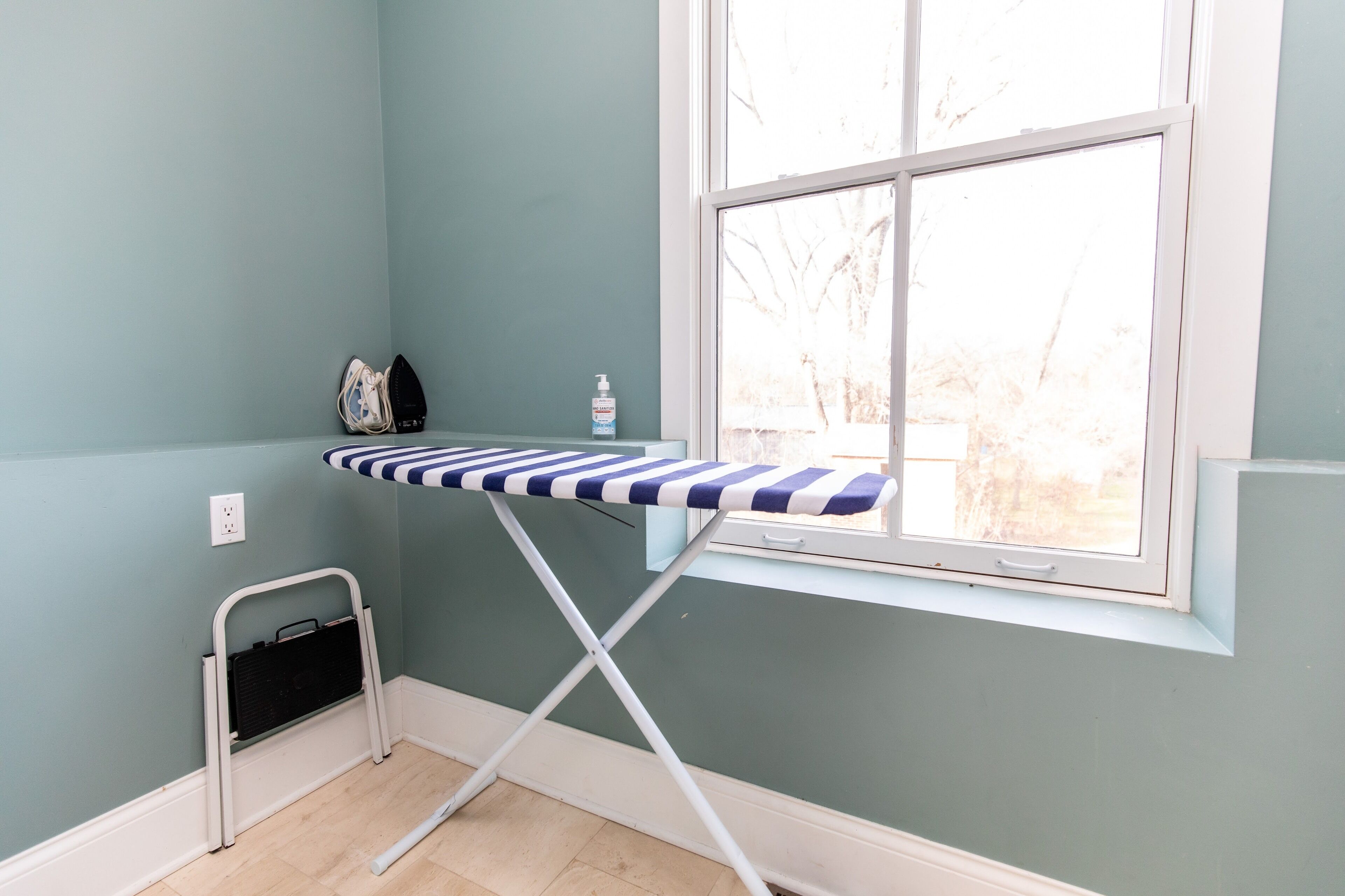 A room with a window, a small ironing board, a basket, and a bottle of hand sanitizer.