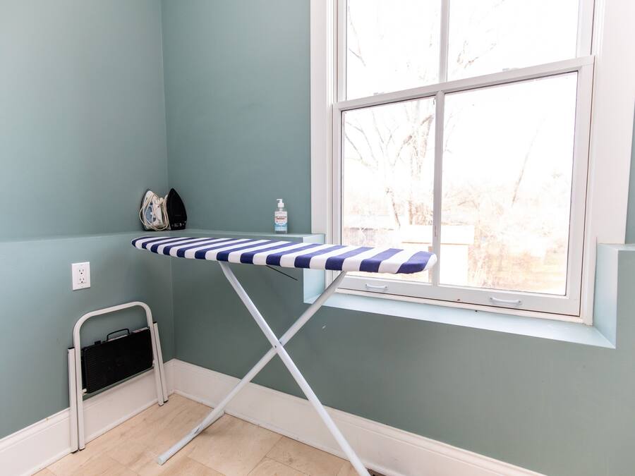 Una habitación con ventana, una plancha pequeña, un cesto y un frasco de desinfectante de manos.