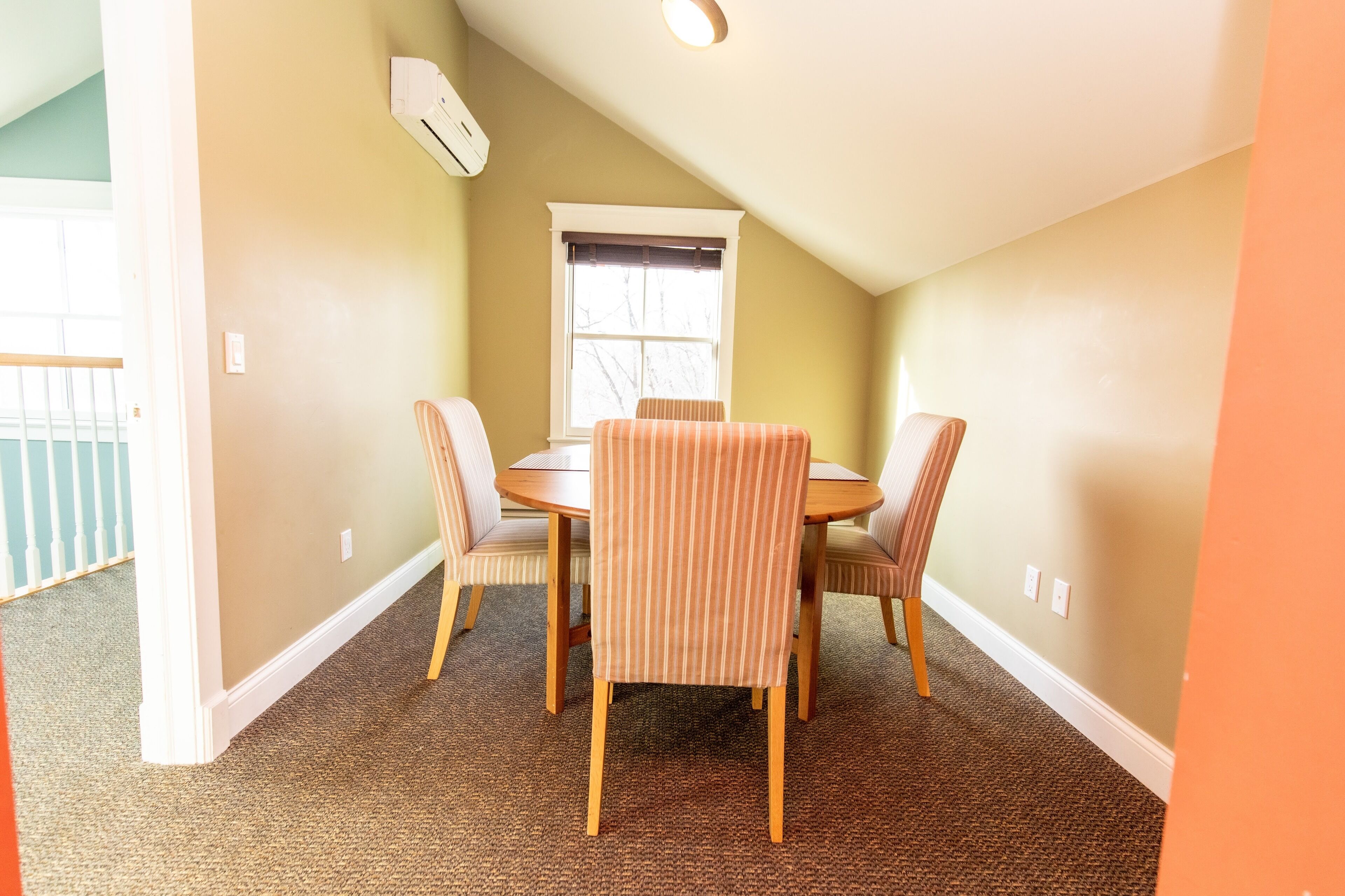 Un comedor con una mesa redonda y cuatro sillas a rayas, una ventana con persianas y un equipo de aire acondicionado en la pared.