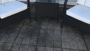 En uteplats med två flätade fåtöljer och ett litet bord, placerad framför ett vitt hus med ett stort fönster.