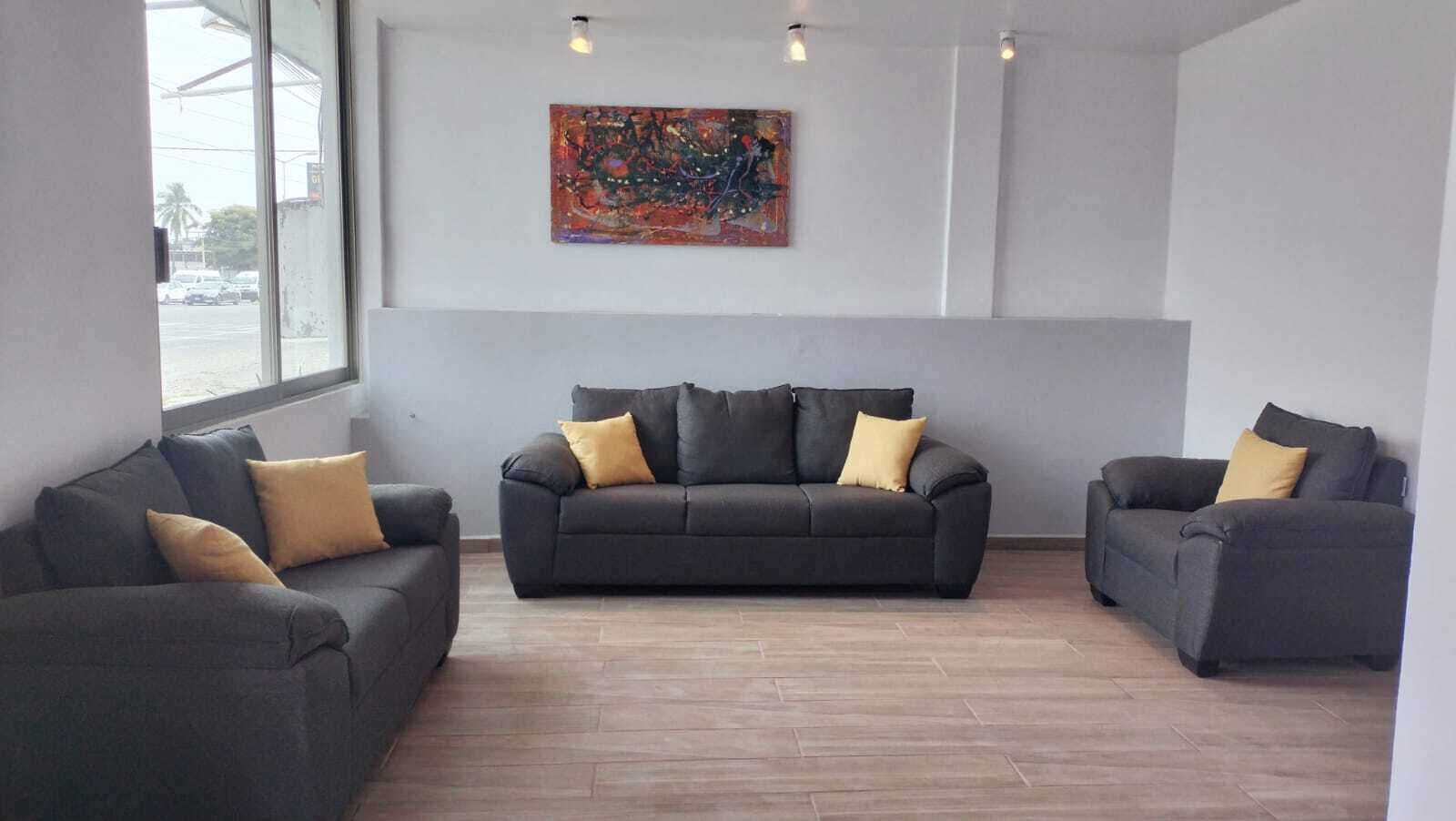 Una sala de estar moderna con tres sofás grises y cojines amarillos, un cuadro abstracto grande en la pared, y un ventanal con vista a la calle.