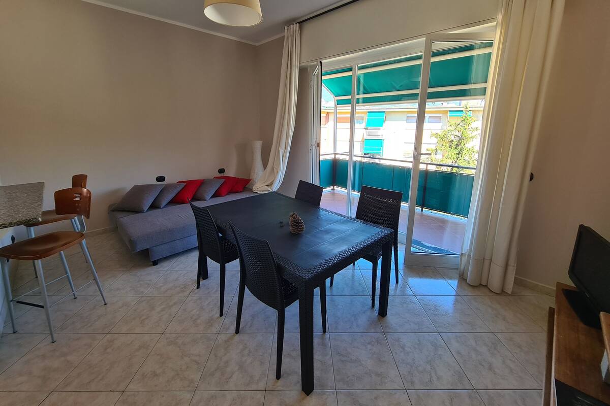 Un salon moderne comprenant une table à manger, des chaises, un canapé avec des coussins, un téléviseur et un balcon avec des rideaux.
