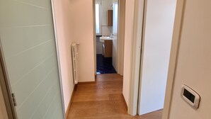 Un couloir doté d’un plancher en bois, de murs blancs et d’une porte donnant sur une salle de bain équipée d’un lavabo et d’un miroir.