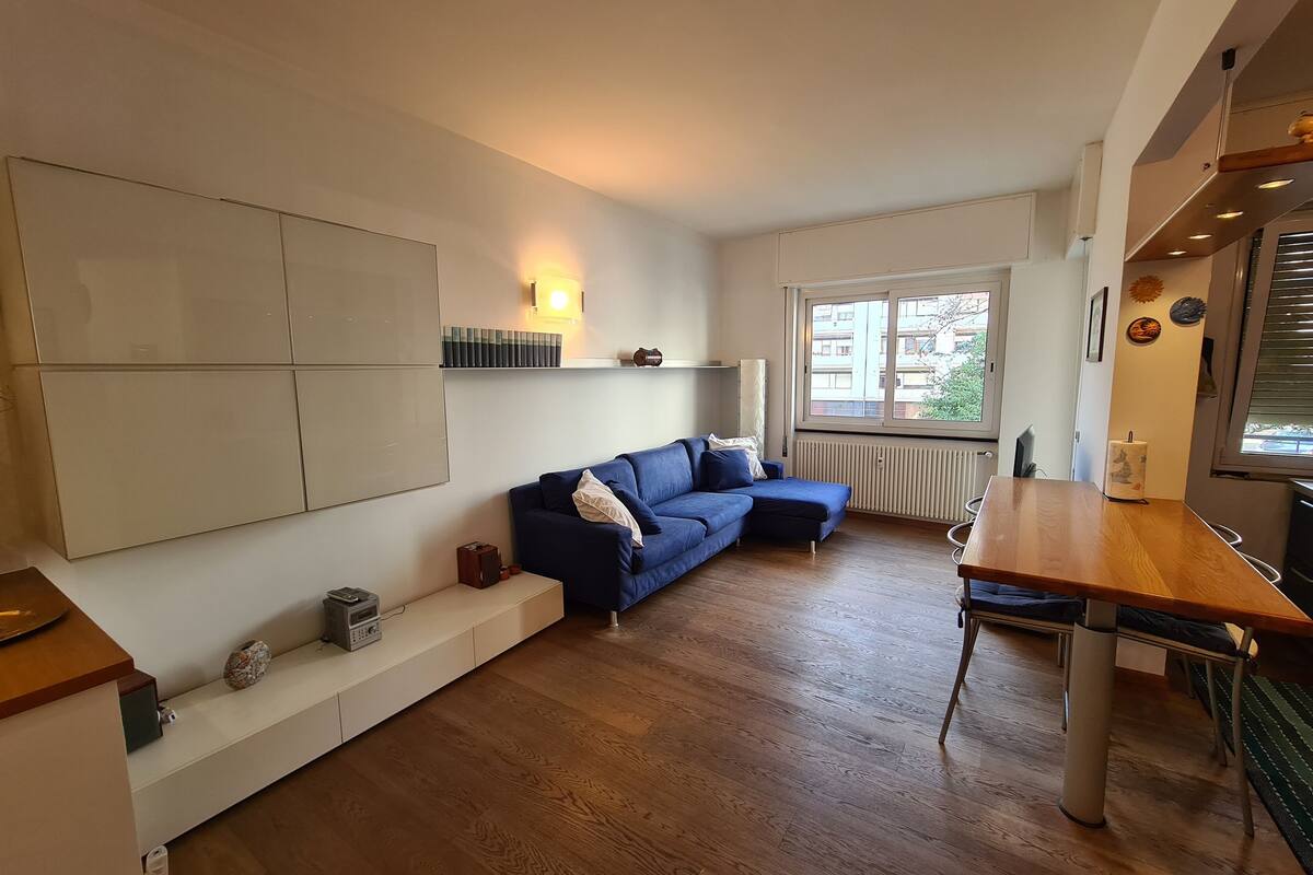 Un salon moderne avec un canapé bleu, une table à manger en bois et une grande fenêtre offrant une vue sur l’extérieur.