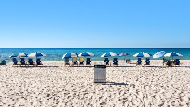 A beach with rows of beach chairs and umbrellas, a signboard, and the ocean in the background. - Emerald Isle 507 The Jolly Roger🌊Beachfront Condo w/ Views + Chair Service (Pensacola Beach)