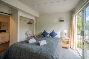 A bedroom with a large bed, bedside tables, a fan, and a view of greenery outside. - Lushingtons Bay - Takamatua (Akaroa)