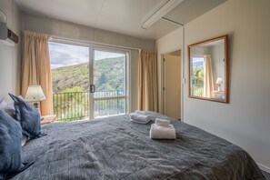 Ein Schlafzimmer mit einem großen Bett, einem Fenster mit Vorhängen, einem Spiegel und Blick auf eine Hügelkuppe.