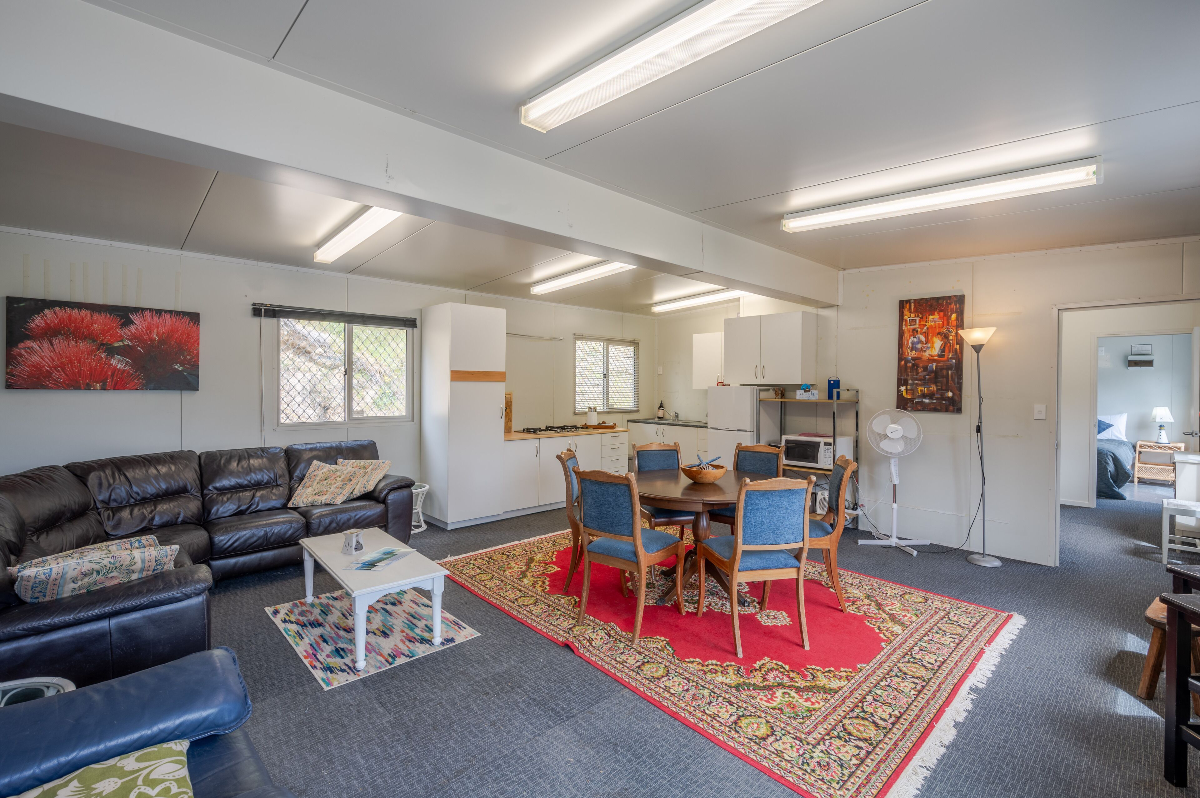 A modern living room with a leather sofa, a dining table with chairs, a red rug, and a kitchen area with cabinets and a refrigerator.