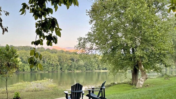 Una terraza de madera con dos sillas Adirondack y una mesa, con vistas a un lago rodeado de árboles y colinas.