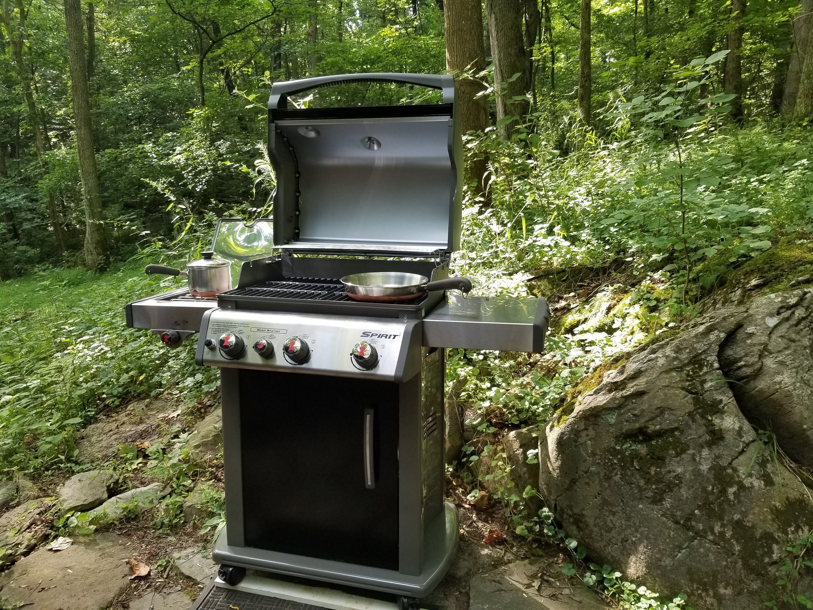 A portable Spirit grill with four burners, a side burner, and a water pan, set up in a forested area.