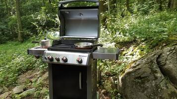 A portable Spirit grill with four burners, a side burner, and a water pan, set up in a forested area.