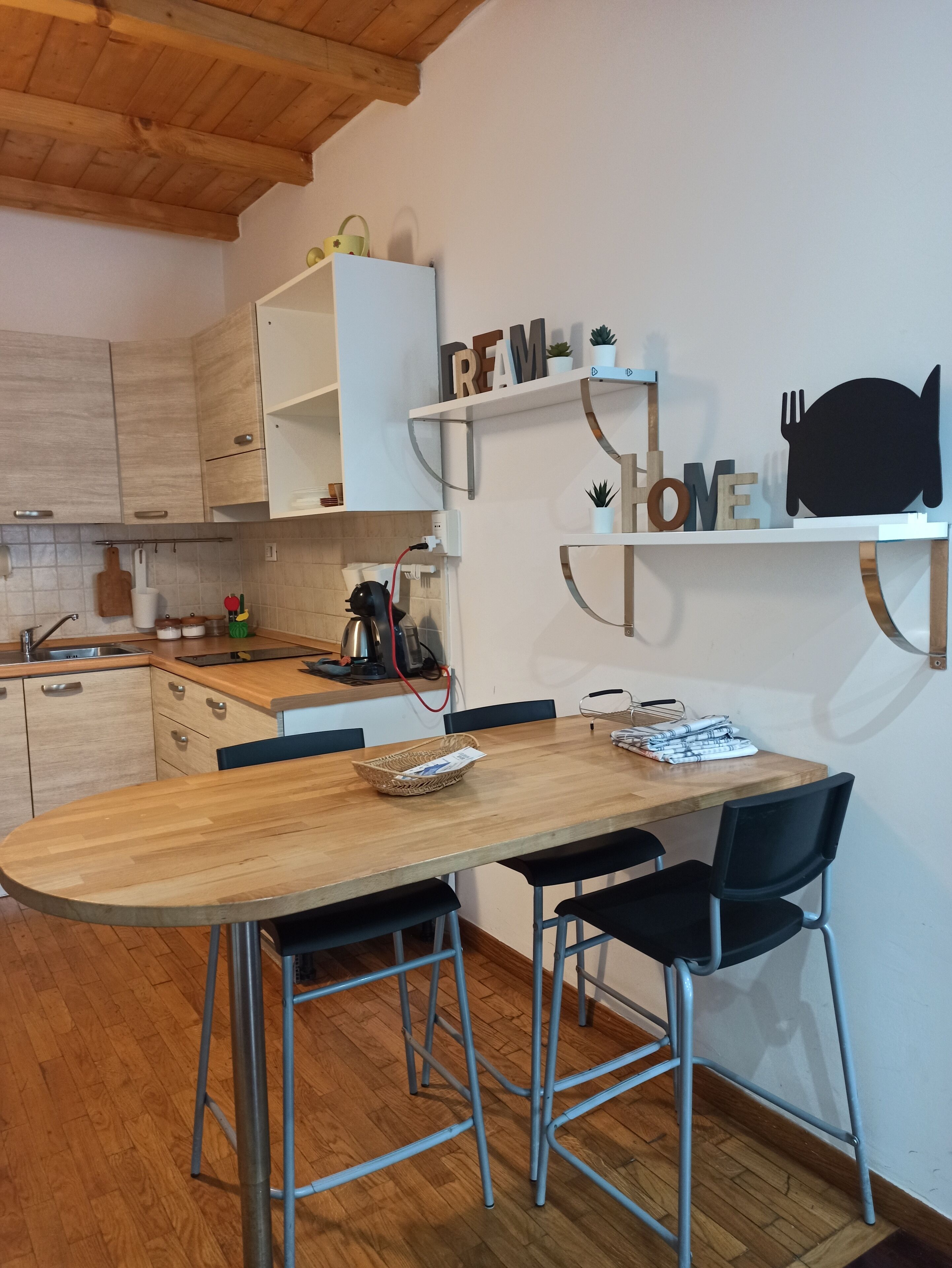 A modern kitchen with wooden cabinets, a wooden dining table, and black chairs. There are shelves with decorative items and a coffee machine on the counter.