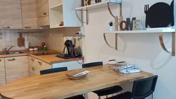 A modern kitchen with wooden cabinets, a wooden dining table, and black chairs. There are shelves with decorative items and a coffee machine on the counter.