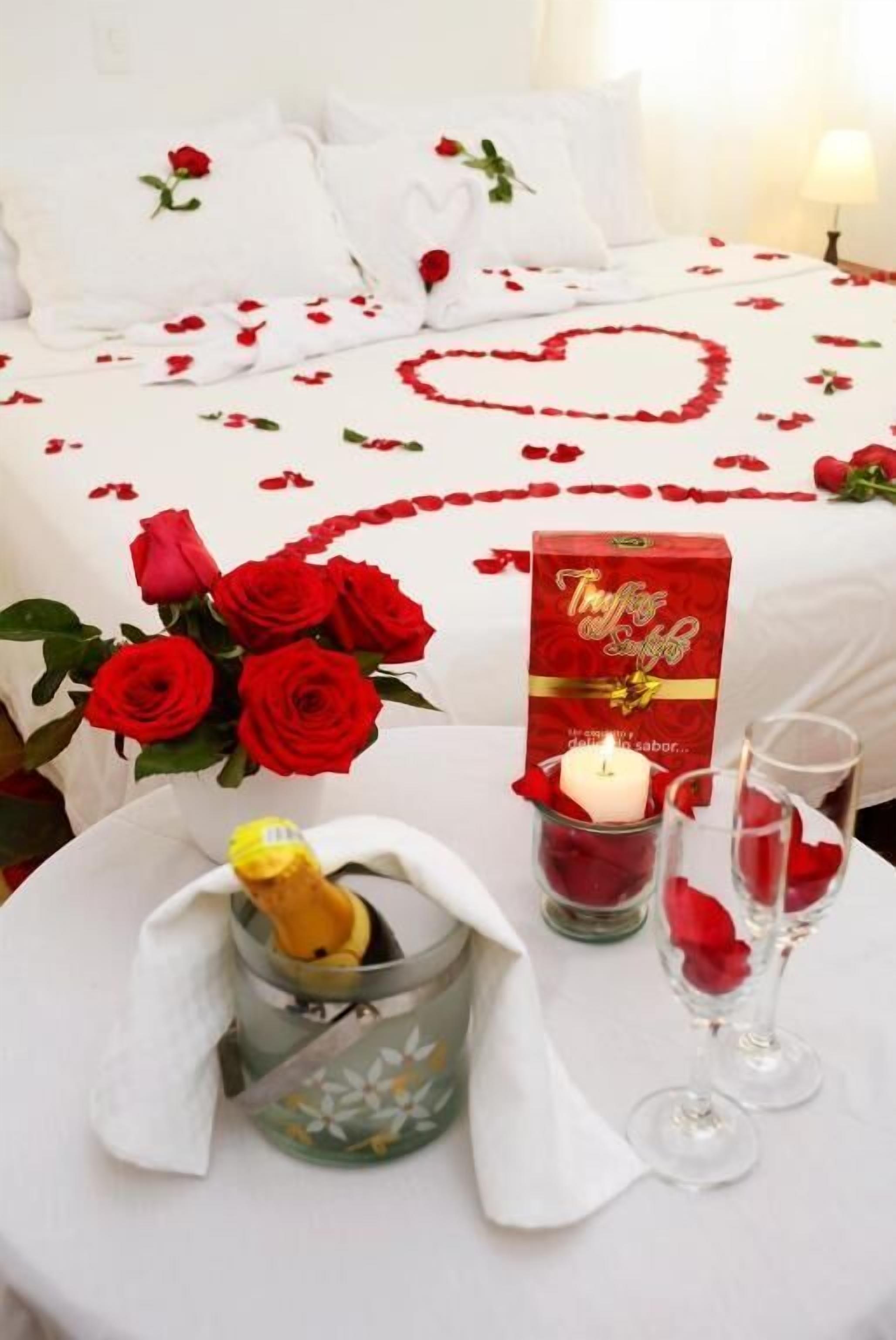 A hotel room with a bed adorned with red rose petals and a heart design, a bouquet of red roses, a bottle of champagne, and two glasses on a round table.