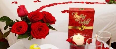 A hotel room with a bed adorned with red rose petals and a heart design, a bouquet of red roses, a bottle of champagne, and two glasses on a round table.