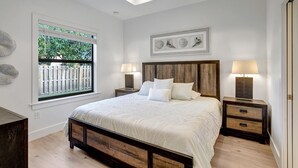 A bedroom with a large bed, two bedside tables with lamps, a wooden headboard, a window with blinds, and a ceiling fan.