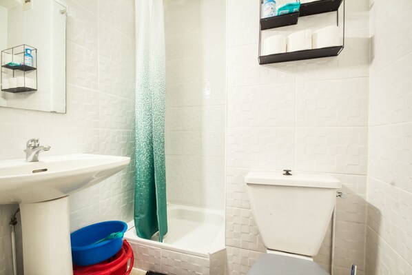 Une salle de bain avec un lavabo, des toilettes et une baignoire, tous de couleur blanche. On y trouve des étagères remplies de produits de toilette et un rideau de douche vert.