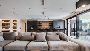 Un salon moderne doté d’un grand canapé d’angle, d’un coin repas avec une table et des chaises, et d’une cuisine avec des meubles sombres et une grande fenêtre.