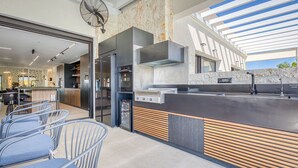 Une cuisine moderne dotée d’une cuisinière intégrée, d’un grand îlot central avec un évier et d’un ventilateur. On peut apercevoir des chaises et un coin bar.