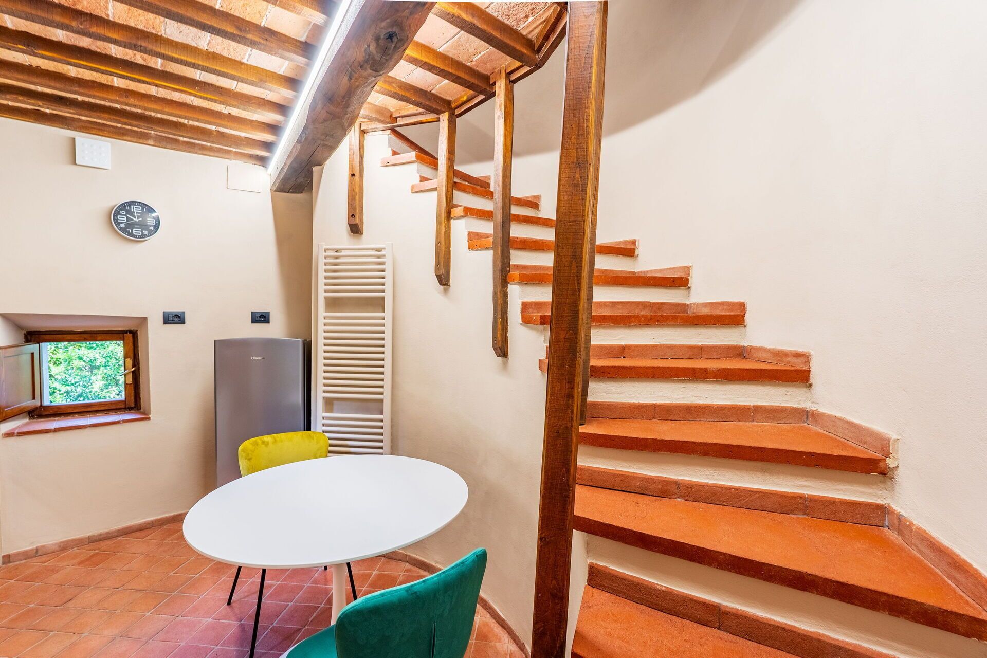 Quarto com uma mesa branca, duas cadeiras, um relógio, um radiador e uma escada.